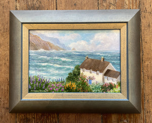 A small painting in a brown wood frame depicting an oceanside cottage scene.  The ocean is a dominant part of the image with small waves rolling in and billowy white clouds in the blue sky.  Rocky cliffs are included in the  upper left hand corner.  There are many colorful flowers around the cottage creating a lovely scene of an oceanfront cottage.  The cottage looks like an English cottage with two chimneys atop a brown roof.  The cottage has five windows showing and a blue front door. 