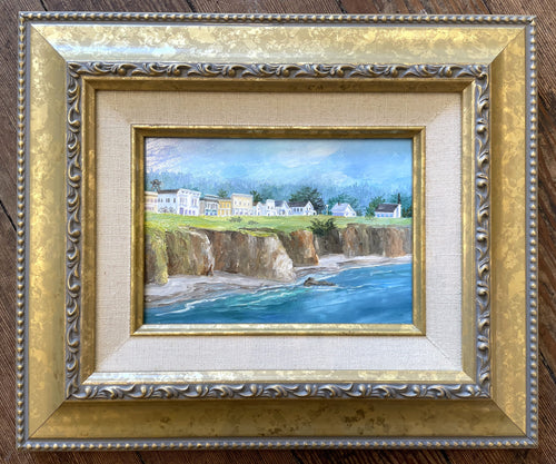 A small painting of the town of Mendocino and the Headlands looking at town from the ocean.  There are a row of houses along Main street including a church and a watertower.  There is green grass atop the cliffs in front of the buildings with ocean and a small strip of beach in the forefront.  There are trees in the background behind the buildings and the sky is blue.  The main colors are blue, green, brown and white with a little yellow.