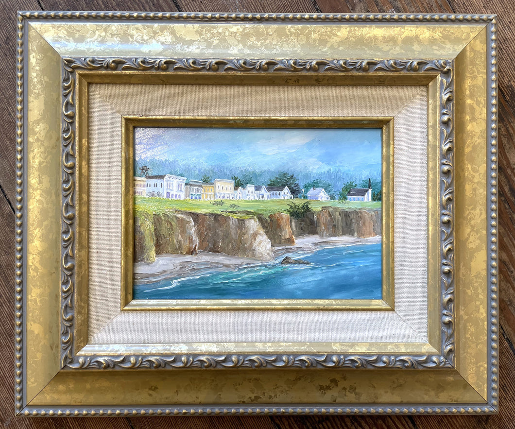 A small painting of the town of Mendocino and the Headlands looking at town from the ocean.  There are a row of houses along Main street including a church and a watertower.  There is green grass atop the cliffs in front of the buildings with ocean and a small strip of beach in the forefront.  There are trees in the background behind the buildings and the sky is blue.  The main colors are blue, green, brown and white with a little yellow.