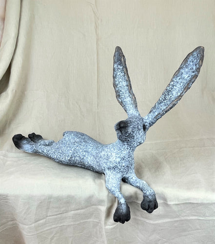 Basil is a bronze sculpture meant to be displayed on a shelf as its two front feet will hang over the edge of the shelf making it look like he is hanging out comfortably there.  He has a blue gray speckled finish with black feet, nose and eyes.  His ears are erect and listening and his feet are kicked out behind him as he is in a laying down position.  