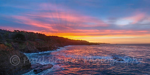 Rays of morning sunrise of gold, salmon and into a blue sky, over the Pacific ocean and cliffs.